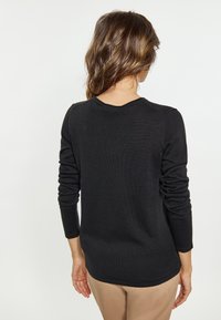 Top negru cu mâneci lungi, cu textură ribbată și decolteu rotund, cu o siluetă ajustată și un tiv ușor curbat; purtat cu pantaloni bej.