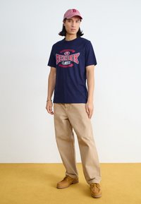 Marineblå bomull T-skjorte med et rødt og hvitt Reebok-logo design, sammen med beige bukser og brune snøresko på et gult gulv.