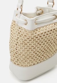 Bolsa de tote tejida con un cuerpo color beige claro, ribete sintético blanco y herrajes en tono dorado. Presenta una superficie texturizada y una forma estructurada.