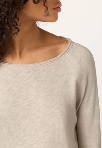 Moteris su garbanotais plaukais dėvi šviesiai smėlio spalvos ilgomis rankovėmis megztinį su neapdorotu apvaliu iškirptės kraštu, žiūrėdama šonu į vientisą foną.