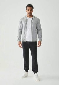 Grauer Reißverschluss-Hoodie mit weißen Kordeln, getragen über einem weißen T-Shirt. Schwarze Jogginghosen und weiße Turnschuhe runden das Outfit ab.