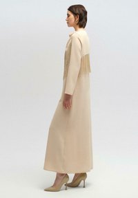Maxi jurk van beige stof met lange mouwen, met een franjen detail op de rug. Draagt bij nude hoge hakken.
