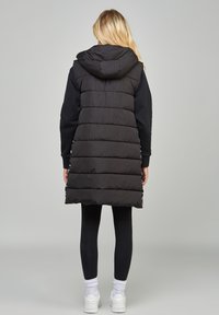 Black puffer vest with a hood, quilted design, and mid-thigh length, worn over a black long-sleeve top and black leggings.