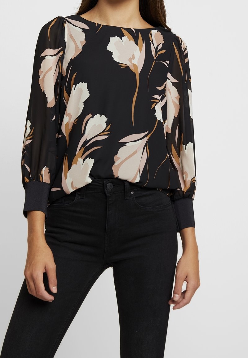 Femme portant un chemisier noir à imprimé floral beige et blanc, et un jean noir taille haute, debout avec les mains détendues le long du corps.