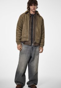 Hnědá bomber bunda z semiše s límcem z ovčí vlny, na sobě světle hnědou košili, doplněnou volnými světle modrými džíny a hnědou obuví.