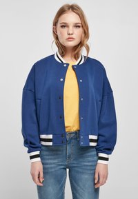 Chaqueta bomber azul con un corte relajado, puños y dobladillo acanalados de rayas blancas y negras en contraste, cerrados con botones a presión plateados. Camiseta blanca visible debajo.