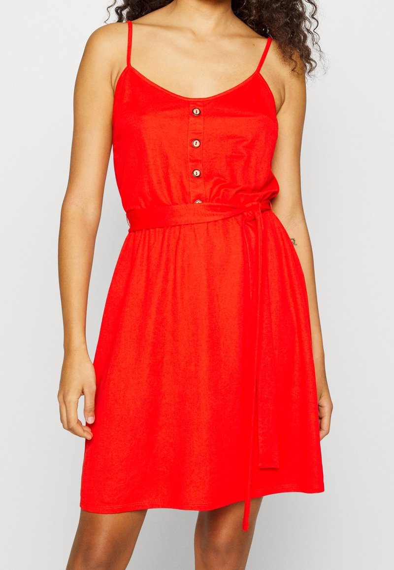 Femme portant une robe rouge sans manches avec des boutons sur la poitrine et une ceinture nouée à la taille sur un fond uni.