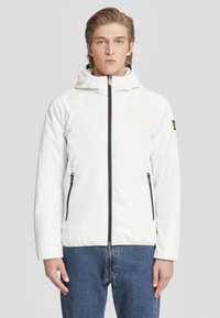 Giacca isolante bianca con chiusura frontale a zip, cappuccio regolabile e due tasche laterali con zip. Presenta rifiniture nere e un logo in evidenza.