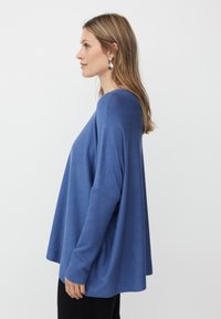 Frau mit langen blonden Haaren, die einen lockeren blauen Pullover und silberne Ohrringe trägt, in Profilansicht vor einem schlichten weißen Hintergrund.