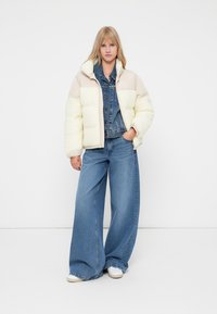 Gevoerde, crèmekleurige jas met een zachte textuur en een aansluitende kraag, gecombineerd met een spijkerjas en wijde blauwe jeans.