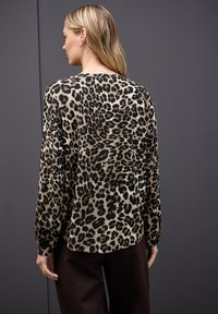 Blůza s leopardím vzorem a dlouhými rukávy, vyrobená z měkké látky, s volným střihem a kulatým výstřihem. Viditelné jsou tmavě hnědé kalhoty.