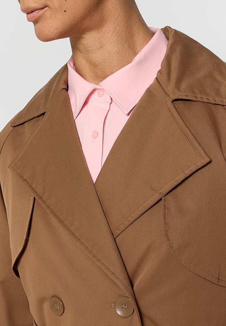 Manteau trench double boutonnage marron avec grands revers, tissu texturé et deux boutons, superposé sur une chemise à col rose pâle.