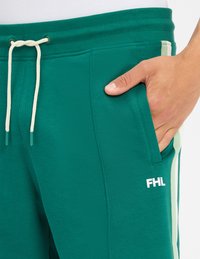 Zielone spodnie sportowe z białym sznurkiem, kieszenią boczną i małym białym logo "FHL" na udzie, noszone przez osobę z ręką w kieszeni.