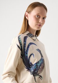 Sudadera beige con un gran gráfico de pavo real en tonos azules y verdes, con un cuello a rayas y un diseño holgado.