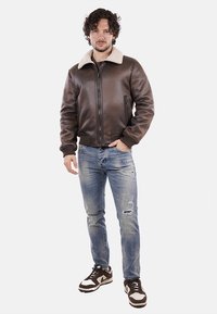 Giacca bomber in pelle marrone con colletto in shearling e chiusura a zip, abbinata a jeans azzurri invecchiati e sneakers scure.