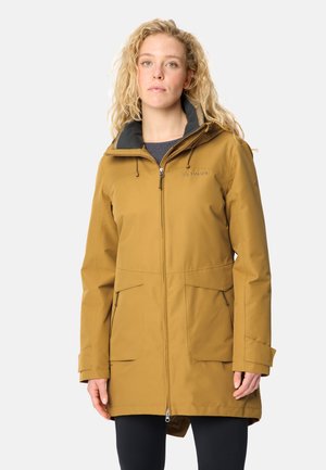 Jonge vrouw met krullend blond haar, gekleed in een mosterdgele capuchonjas met voorkantenzakken en een zwarte broek, staand tegen een effen achtergrond.