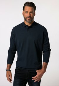 Polo de manga larga azul marino con cuello, tapeta de tres botones y un sutil logo en relieve en el pecho. Se lleva con jeans oscuros.