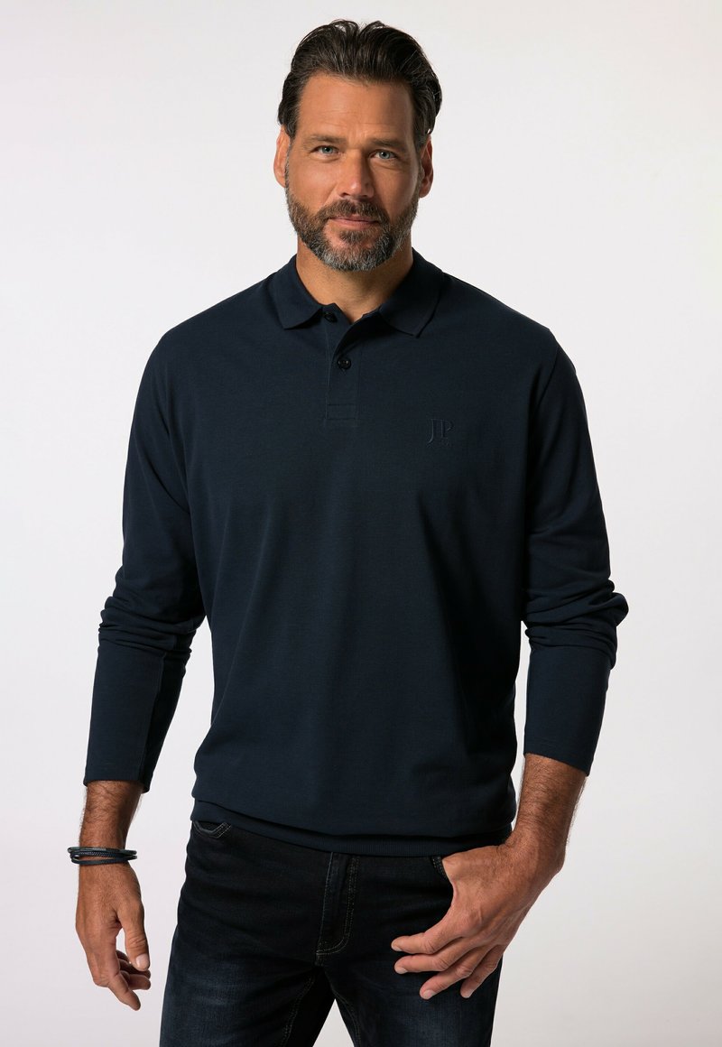 Polo de manga larga azul marino con cuello, tapeta de tres botones y un sutil logo en relieve en el pecho. Se lleva con jeans oscuros.