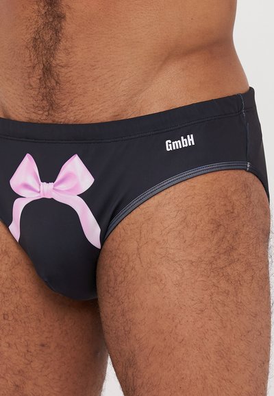 Maillot de bain noir avec un grand graphique de nœud rose à l'avant, et un logo "GmbH" sur le côté droit. Texture du tissu lisse.