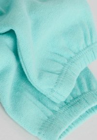 Turkoosinsininen neulottu kangas, jossa on pehmeä rakenne, kerätyt hihansuut ja sileä reunasauma, korostaen vaaleaa ja ilmavaa muotoilua.