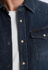 Ciemnoniebieska koszula dżinsowa z kołnierzykiem zapinanym na guziki, z żółtymi i czarnymi metalowymi zatrzaskami, kieszenią na piersi oraz subtelnymi detalami w szwach.