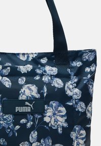 Niebieska torba typu tote z błyszczącym wykończeniem. Posiada wzory białych i jasnoniebieskich kwiatów oraz niebieską metkę z logo "PUMA" w jasnoszarym kolorze.