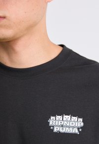 Nærbillede af en person iført en sort skjorte med et lille logo af fem hvide kattehoveder over teksten "RIPNDIP PUMA" på brystet.