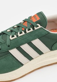 Zelené semišové a síťovinové tenisky s bílými koženými pruhy, texturovanými tkaničkami a oranžovým logem Adidas na jazyku. Světlá gumová podešev.