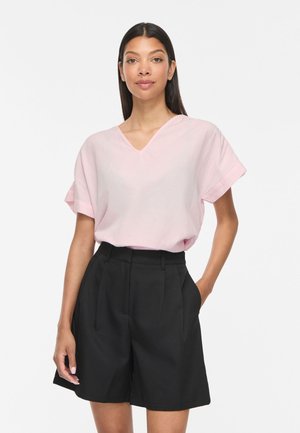 Femme aux longs cheveux foncés portant un haut rose clair à manches courtes et col en V, ainsi qu’un short noir taille haute sur mesure, debout avec une main dans la poche.