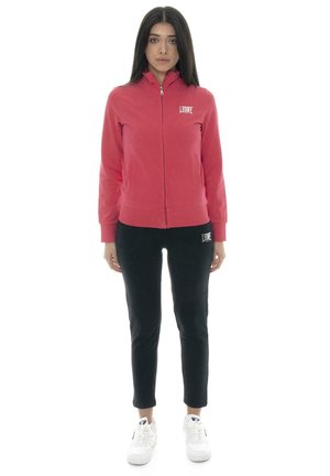 Une femme porte une veste rose zippée, un pantalon noir et des baskets blanches sur un fond blanc uni.