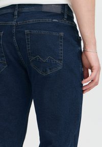 Mørkeblå denimjeans har to baklommer med karakteristiske trekantede sømdetaljer. Stoffet føles jevnt med en svak glans.