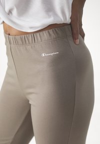 Brązowe, elastyczne legginsy z gumką w pasie. Zawierają małe, białe logo "Champion" po boku. Gładka faktura, dopasowany krój.