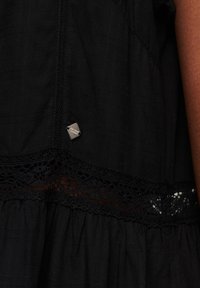 Svart tygklänning med texturerade ränder, prydd med spetsdetaljer längs fållen och en liten metallisk logotagg på sidan.