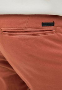 Rust-colored pants with a smooth texture, featuring a back pocket and a small black label with "JACK & JONES" stitched on it.