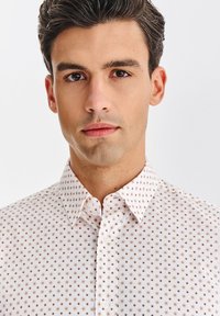 Biała koszula z drobnym brązowym wzorem w kropki, z kołnierzykiem zapinanym na guziki i gładką bawełnianą fakturą, prezentowana na modelu męskim.