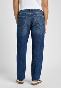 Vaqueros de mezclilla azul con un corte relajado, que cuentan con bolsillos traseros, un corte recto y costuras visibles. Llevados con zapatos de color claro.