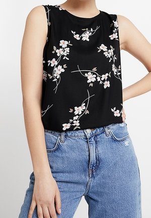 Czarny top bez rękawów z białym i różowym kwiatowym wzorem, noszony z jasnoniebieskimi wysokim stanem dżinsami, ręka oparta na biodrze.