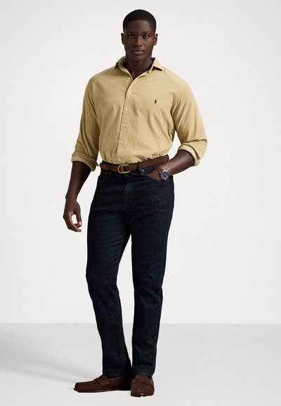 Homem vestido com uma camisa de manga longa bege, jeans escuros, cinto castanho, mocassins castanhos e um relógio de pulso, de pé com uma mão no bolso contra um fundo simples.