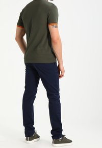 Mörkgrön pikétröja med orange detaljer på ärmarna, tillsammans med marinblå byxor och grå sneakers, sedd bakifrån.