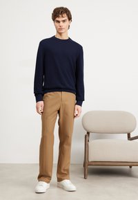Pullover a maglia blu navy, pantaloni beige e sneaker bianche. Il modello è appoggiato a uno sfondo neutro. Design semplice e minimalista senza motivi.