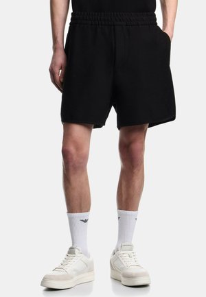 Mand iført sorte shorts med elastisk talje, hvide ankelsokker med sort logo og off-white sneakers, mod en ensfarvet baggrund.