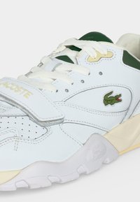 Hvid Lacoste sneaker med grønne detaljer, hvide snørebånd, velcrolukning og en grøn krokodillelogo på siden.