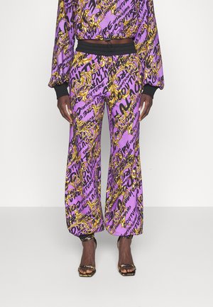 Lila Hose mit schwarzer Schrift und goldenen Barockmustern, elastischem Bund und Knöchelabschlüssen, kombiniert mit passendem Oberteil und offenen Pumps.