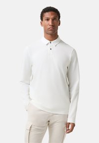 Mann in einem weißen langärmeligen Poloshirt und hellbeigen Cargo-Hosen, der vor einem schlichten hellen Hintergrund steht.
