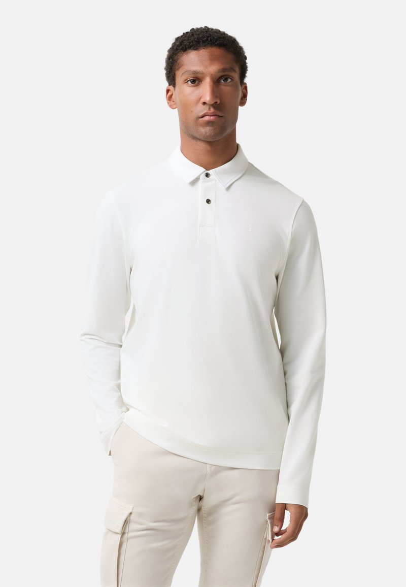 Mann in einem weißen langärmeligen Poloshirt und hellbeigen Cargo-Hosen, der vor einem schlichten hellen Hintergrund steht.