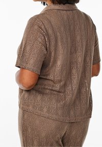 Brązowa bluzka z krótkim rękawem, wykonana z perforowanej tkaniny z diamentowym wzorem i kołnierzykiem. Widok z tyłu, ukazujący teksturowe detale.