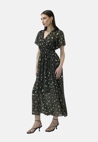 Robe maxi en tissu transparent noir avec un motif floral doré. Col en V, manches courtes, taille élastique, jupe fluide. Portée avec des talons noirs à lanières.