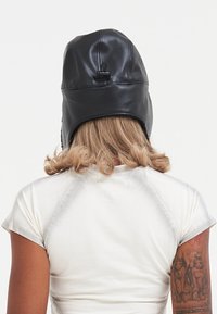 Cappello da aviatore in pelle nera con una texture liscia, dettagli cuciti e una cintura regolabile sul retro, indossato da una persona con capelli mossi.