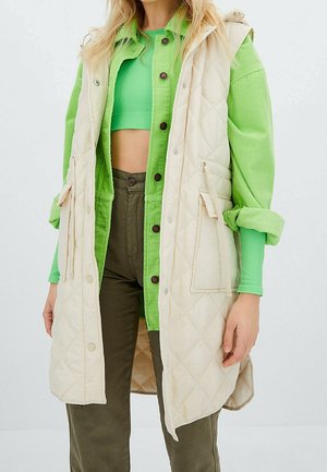 Kvinde iført en beige quiltet ærmeløs vest over en grøn cropped top, grøn overshirt og olivengrønne bukser mod en hvid baggrund.