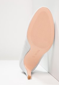 Semelle d'une chaussure à talons hauts blanche avec grip texturé et petit talon, positionnée à la verticale contre un mur d'angle blanc.